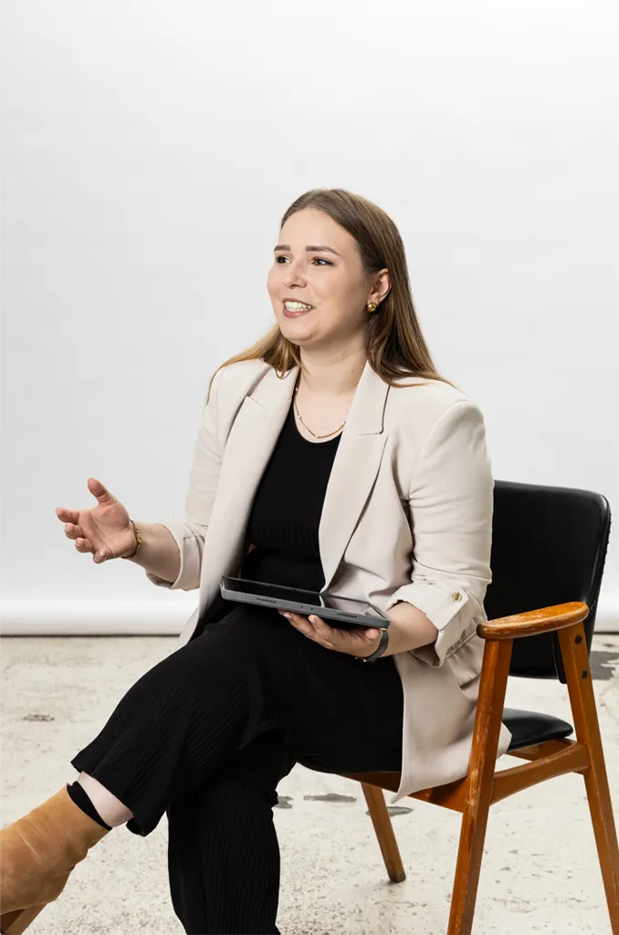 Porträt von Maren Dinges (Content mit Maren), vor weißem Hintergrund sitzend und offen gestikulierend, mit Tablet in der Hand und beigem Blazer.